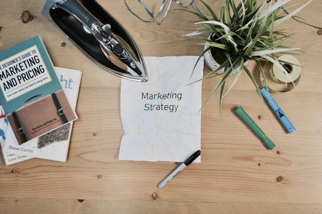 A imagem mostra uma mesa com canetas, livros, uma planta e um papel com os escritos "marketing strategy"